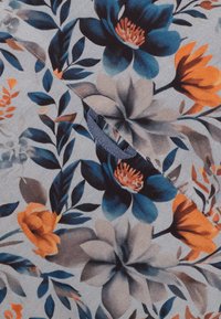 Bloemstof met grote blauwe, oranje en beige bloemen en bladeren op een lichtgrijze achtergrond met zichtbare stiksels en een stoffen lus.