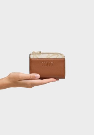 Mano sosteniendo una pequeña cartera con cremallera de color marrón y beige con el nombre de la marca "TOUS" grabado en el frente.