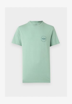 Homme aux cheveux courts et foncés portant un t-shirt vert clair Rip Curl Surf Co et un pantalon blanc, vu de dos sur un fond uni.