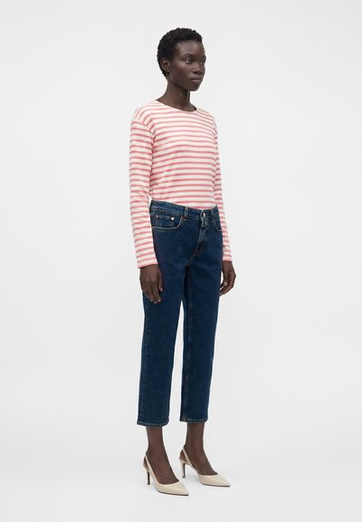 Femme debout portant un haut à manches longues rayé rose et blanc, un jean taille haute bleu foncé et des talons hauts beiges pointus.