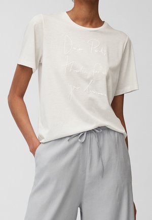 Personne portant un t-shirt blanc avec un texte en cursive "Cher Parent, merci pour toutes tes leçons" et un pantalon gris clair à cordon de serrage.