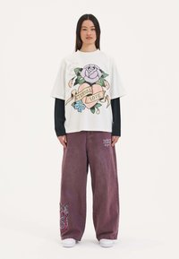 Witte oversized t-shirt met een bloemdesign met een roze roos en een lint waarop "ETERNAL LOVE" staat. Combinatie met paarse wijde broek.