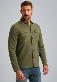 Olijfgroene knoopshirt met een gestructureerde stof, twee borstzakken en lange mouwen. Het materiaal lijkt lichtgewicht en ademend te zijn.