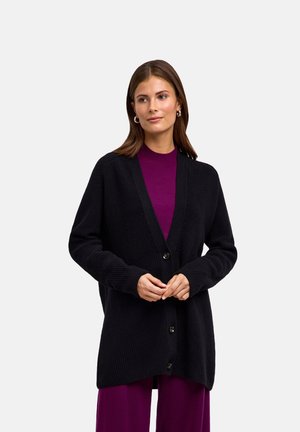 Kobieta ubrana w czarny zapinany na guziki kardigan na fioletowej bluzce i spodniach, stoi ze splecionymi rękami, spogląda lekko na bok na jednolitym tle.
