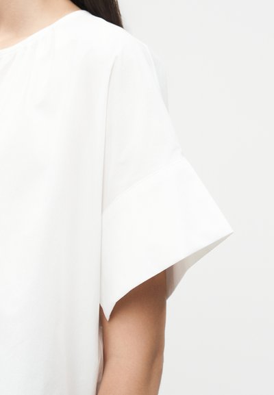 Γυναίκα που φοράει λευκό τοπ με κοντά μανίκια, φαρδιές, κεκλιμένες μανσέτες και στρογγυλή λαιμόκοψη σε απλό ανοιχτό φόντο.