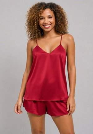 Femme aux cheveux bouclés souriante, portant un haut caraco en satin rouge et un short assorti sur fond gris.