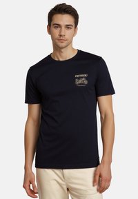 Camiseta de algodón azul marino con cuello redondo, mangas cortas y un logotipo impreso en dorado con diseño de motocicleta en la parte superior izquierda del pecho.