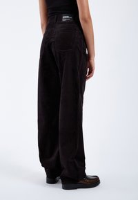 Pantalones de pana anchos en negro con costuras prominentes, bolsillos traseros y una etiqueta en la cintura. La textura es acanalada y suave.