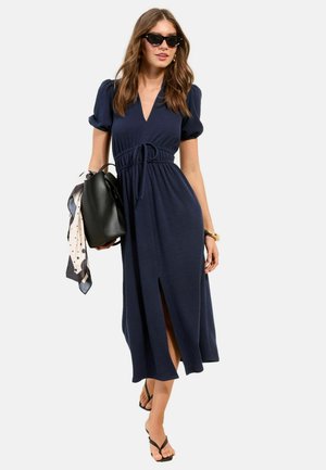 Frau trägt ein marineblaues Midikleid mit Puffärmeln und vorderem Schlitz, hält eine schwarze Handtasche mit gemustertem Schal, trägt schwarze Sandalen und Sonnenbrille.