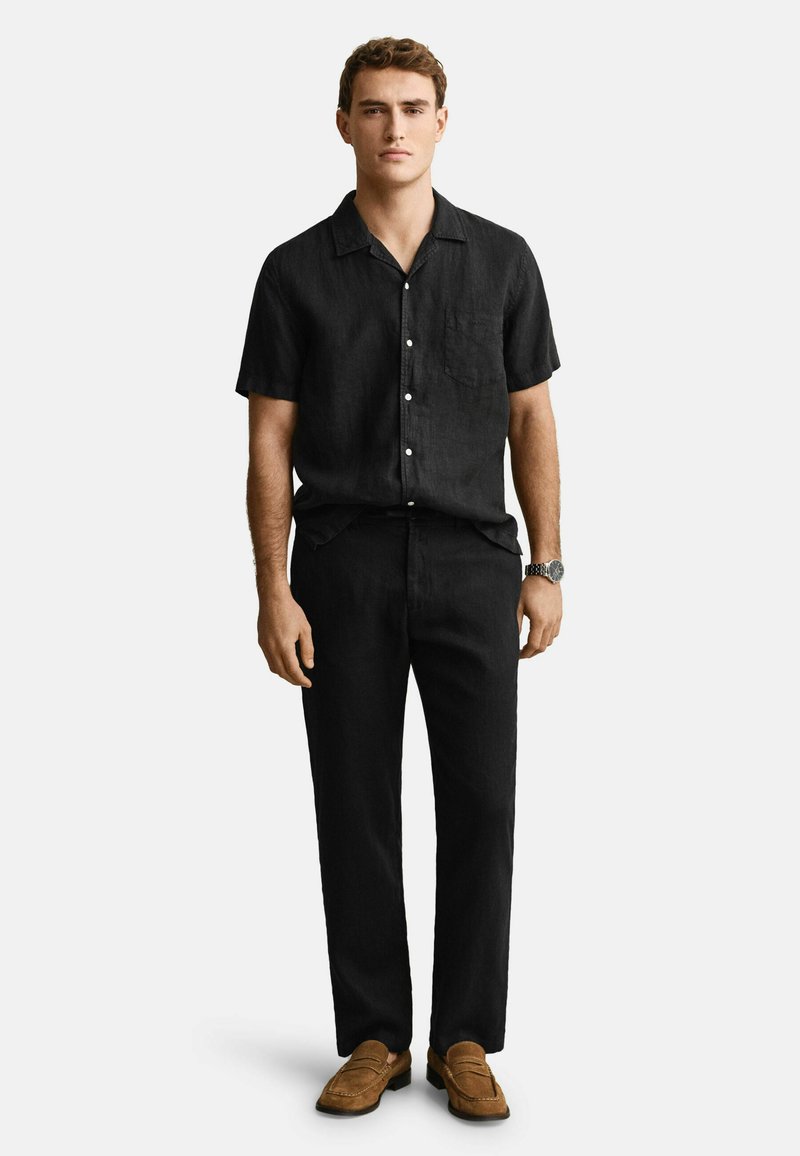 Junger Mann stehend, trägt ein schwarzes kurzärmeliges Hemd mit Knöpfen, schwarze Hose, braune Loafers und eine Armbanduhr am linken Handgelenk.