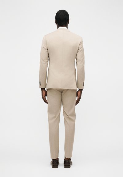 Άνδρας που στέκεται με την πλάτη, φορώντας μπεζ κοστούμι με ταιριαστά παντελόνια και σκούρα καφέ επίσημα παπούτσια σε λευκό απλό φόντο.