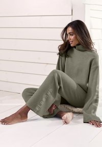 Olivenfarbenes Loungewear-Set mit Rollkragen und weitem Bein. Weicher Stoff, lockere Passform und Logodetail am Hosenbein.