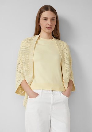 Femme aux longs cheveux bruns portant un châle en maille jaune clair sur un pull jaune pâle et un pantalon blanc, debout les mains dans les poches.