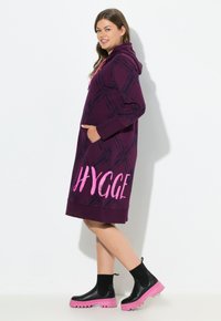Abito felpa viola con cappuccio e motivo geometrico nero, dotato di tasche frontali e la scritta "HYGGE" in rosa. Indossato con stivali neri alla caviglia con suola rosa.