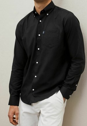 Homme portant une chemise noire à boutons blancs et un pantalon blanc, debout contre un mur clair, une main dans la poche.