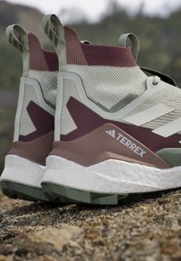 Kevyet vaelluskengät vaalean vihreässä ja maroonissa, joissa on teksturoitu päällinen, joustava muotoilu ja pehmustettu valkoinen välipohja vihreillä yksityiskohdilla.