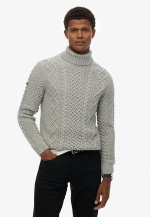 Suéter de cuello alto de punto trenzado en gris claro con patrones texturizados, puños y dobladillo acanalados; llevado sobre una camisa blanca y combinado con pantalones oscuros.