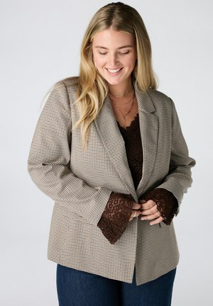Glimlachende blonde vrouw die een geruit blazer draagt over een bruine kanten top en blauwe jeans, ze past haar blazer met beide handen aan.