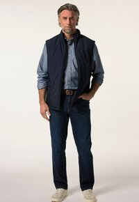 Marinblå vadderad väst över en denimskjorta, tillsammans med blå jeans och ljusa sneakers; har en hög krage och sidofickor.