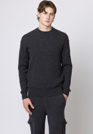 Giovane uomo dalla pelle chiara e capelli castani, indossa un maglione grigio scuro a trecce e pantaloni abbinati, in piedi con una mano in tasca.