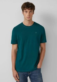 Junger Mann mit lockigem braunem Haar, der ein türkisfarbenes T-Shirt mit kurzen Ärmeln und blaue Jeans trägt und vor einer schlichten Wand steht.