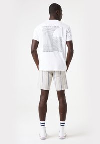 Camisa de algodón blanca con un diseño de arte en línea negra en la espalda. Combinada con pantalones cortos rayados en beige y zapatillas blancas con acentos azules.