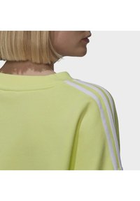 Světlý žlutý svetr s jemnou texturou, který má na rameni tři bílé pruhy a klasický kulatý výstřih.