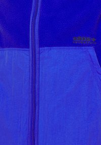 Chaqueta azul real con cremallera que presenta un tejido superior texturizado y un tejido inferior liso, adornada con un logo dorado de "adidas Adventure".