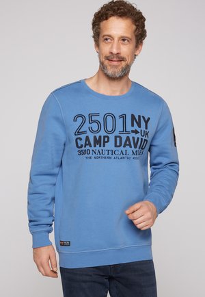 Sudadera azul con cuello redondo, mangas largas y texto impreso en negro. Cuenta con una etiqueta en el dobladillo inferior y puños acanalados.