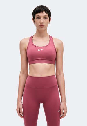 Kobieta z krótkimi ciemnymi włosami, ubrana w dopasowany różowy stanik sportowy i legginsy z wysokim stanem z białymi logo Nike, stojąca na jednolitym tle.