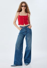 Rotes, kurzes Tanktop mit Knopfdetails, kombiniert mit hochTaillierten, weit geschnittenen blauen Jeans. Das Outfit wird durch spitze Stiefel ergänzt.