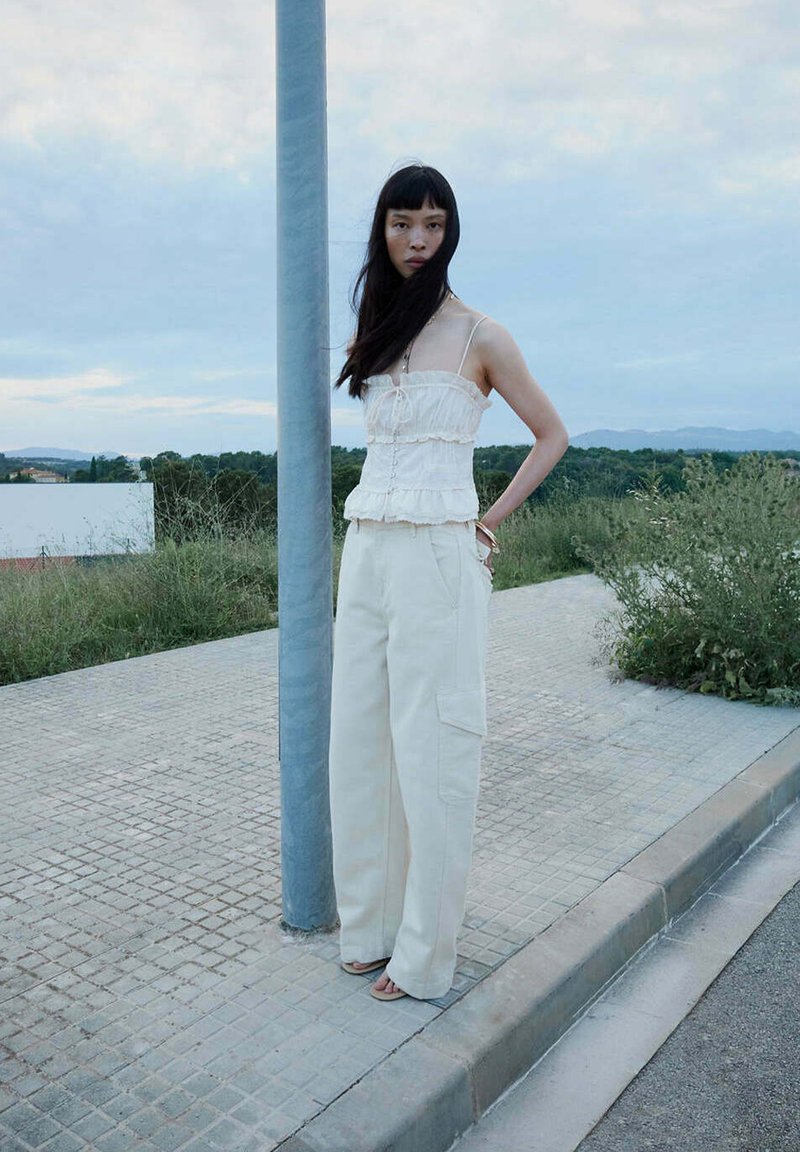 Femme aux longs cheveux foncés portant un haut blanc sans manches et un pantalon blanc large, debout à côté d’un lampadaire sur un trottoir près de la verdure.