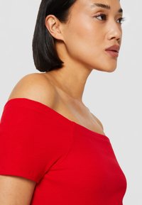 Top rosso a coste con scollo off-the-shoulder e maniche corte. La superficie liscia e il design aderente mettono in risalto il collo e le spalle.