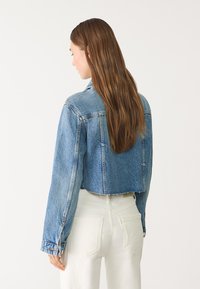 Skrajšana denim jakna v svetlo modri barvi, z dolgimi rokavi, klasičnim ovratnikom in minimalnim strganjem na robu. Prikazana s hrbtne strani.