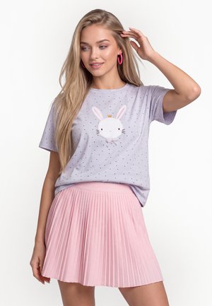 Blondynka z długimi włosami, nosząca różowe kolczyki koła, jasnofioletowy t-shirt z grafiką królika oraz różową plisowaną spódnicę.