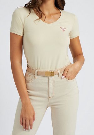 Kobieta ubrana w beżowy T-shirt z dekoltem w serek z małym logo Guess na piersi oraz pasujące beżowe spodnie z klamrą Guess, ręka w kieszeni.