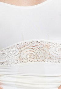 White bodysuit featuring a lace panel with floral and swirled patterns at the waist. Smooth texture, fitted design, and stretchy material.