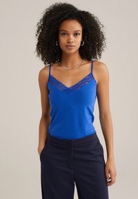 Blå camisole-topp med spetsdetaljer vid halslinjen, med tunna justerbara axelband. Kombinerad med mörkblå byxor. Slät textur.