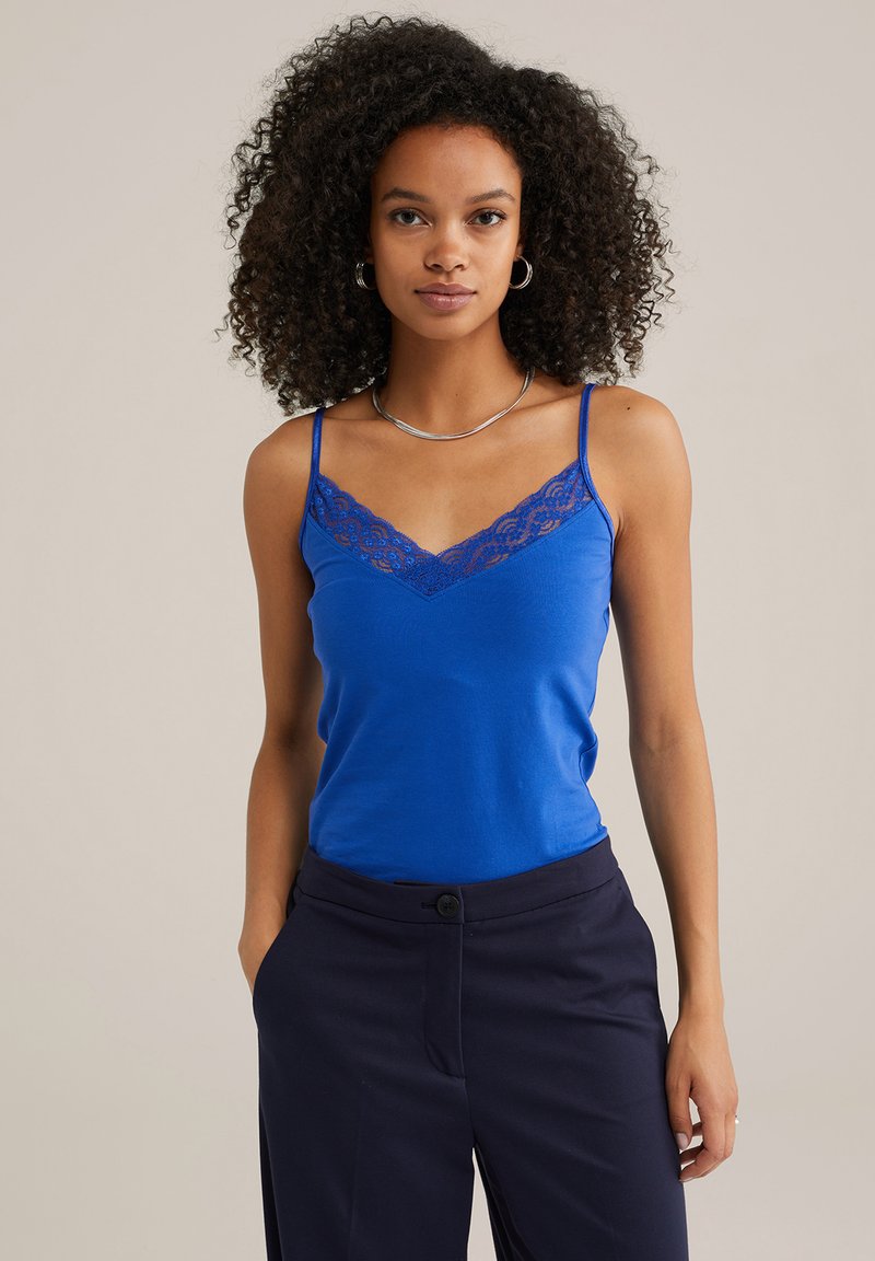 Blå camisole-topp med spetsdetaljer vid halslinjen, med tunna justerbara axelband. Kombinerad med mörkblå byxor. Slät textur.