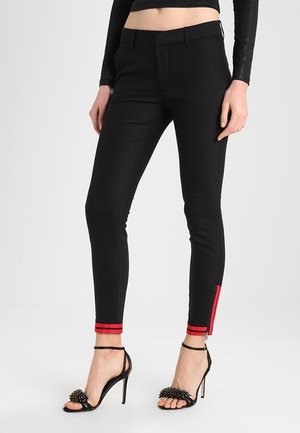 Schmale schwarze Hose mit roten Streifendetails am Knöchel, getragen mit schwarzen Sandalen mit hohen Absätzen und dekorativen Nieten.