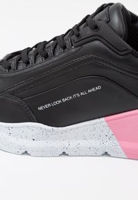 Sneakers noirs en cuir texturé, avec une semelle blanche tachetée et un accent rose. Le texte sur le côté indique : "NE JAMAIS REGARDER EN ARRIÈRE, TOUT EST DEVANT."
