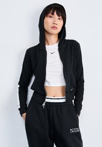 Camisola preta com fecho e capuz, apresentando riscas brancas nas laterais. Usada sobre uma t-shirt branca curta e combinada com calças de treino pretas com o logótipo "NIKE".
