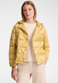 Giacca trapuntata gialla con cappuccio, caratterizzata da una finitura lucida e chiusura con bottoni a pressione, indossata sopra un maglione lavorato a trecce color crema e pantaloni beige.