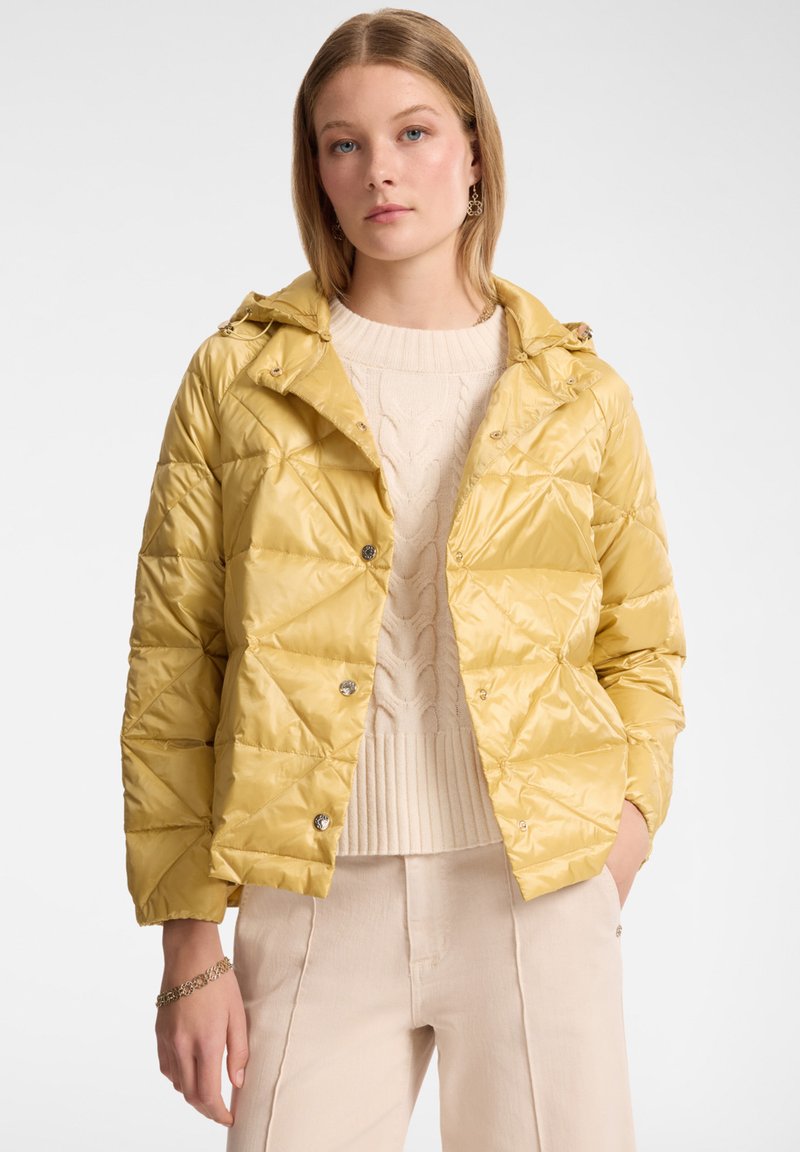Giacca trapuntata gialla con cappuccio, caratterizzata da una finitura lucida e chiusura con bottoni a pressione, indossata sopra un maglione lavorato a trecce color crema e pantaloni beige.