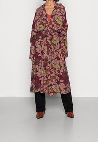 Lange bloemenjurk in roze en aardetinten, met een strik taille en brede mouwen. Gekozen boven een rode top en zwarte broek, met bruine haklaarzen.