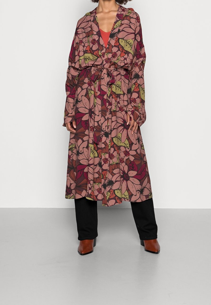 Lange bloemenjurk in roze en aardetinten, met een strik taille en brede mouwen. Gekozen boven een rode top en zwarte broek, met bruine haklaarzen.