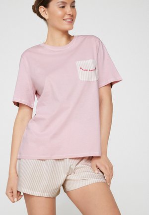 Frau trägt ein pinkfarbenes Kurzarmshirt mit gestreifter Tasche mit der Aufschrift "sweet dreams" und passenden gestreiften Shorts, lächelt leicht und schaut nach rechts.