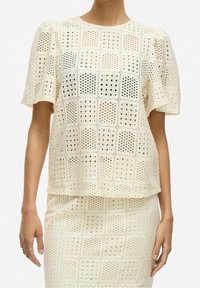 Kvinna som bär en krämfärgad virkad topp och matchande kjol med rutigt mönster och korta ärmar, med händerna avslappnat vid sidorna.