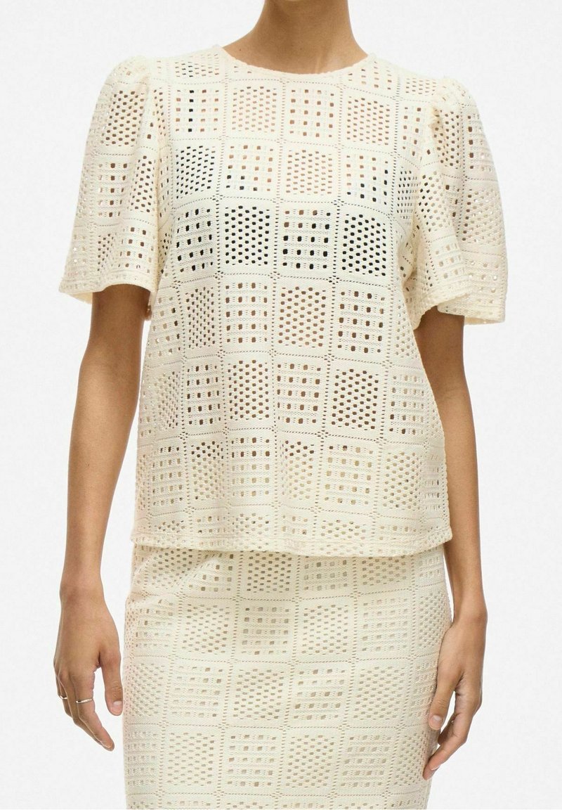 Kvinna som bär en krämfärgad virkad topp och matchande kjol med rutigt mönster och korta ärmar, med händerna avslappnat vid sidorna.