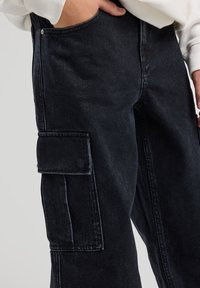 Pantaloni cargo in denim neri con tasca laterale, cuciture a contrasto e vestibilità rilassata. Il tessuto presenta una texture leggermente consumata.